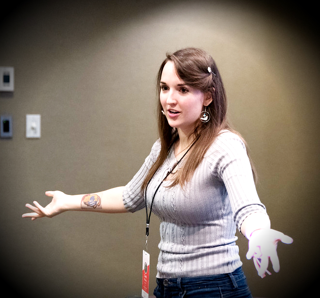 Sarah Snow, a white woman with medium brown hair and brown eyes wearing a grey sweater, makes a shrug movement.